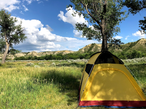 A yellow tent set up in a grassy field, surrounded by trees and hills under a blue sky with fluffy clouds.