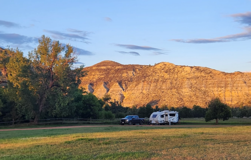 A camper and truck parked on green grass with mountains in the background under a clear sky at sunset.