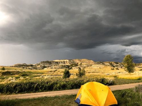 A yellow tent in the foreground with dark storm clouds looming over a hilly landscape.