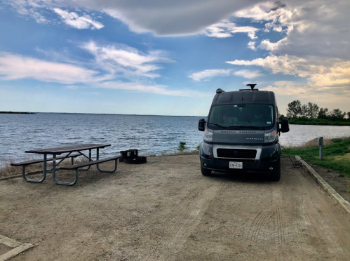 A camper van parked by a lake with a picnic table nearby under a partly cloudy sky.