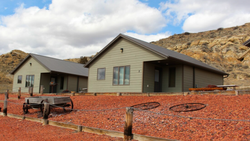 Two modern cabins with a gravel landscape, surrounded by hills and a cloudy sky, featuring rustic wagon wheels in the foreground.