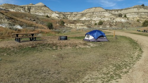 A blue tent set up at a campsite near a picnic table and fire pit, surrounded by rocky hills and clear blue skies.