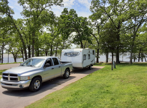 A silver pickup truck towing a white camper trailer parked near a lake, surrounded by green trees and blue sky.