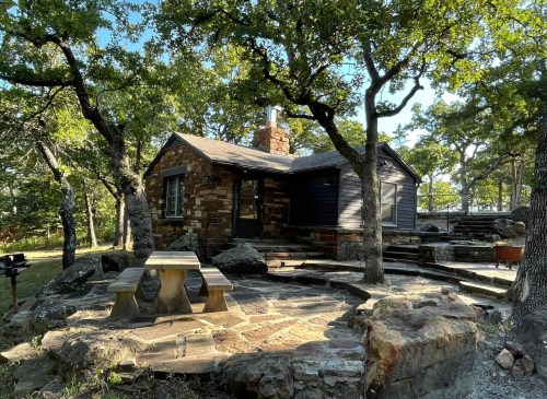 A rustic stone cabin surrounded by trees, featuring a picnic table and a stone pathway in a natural setting.