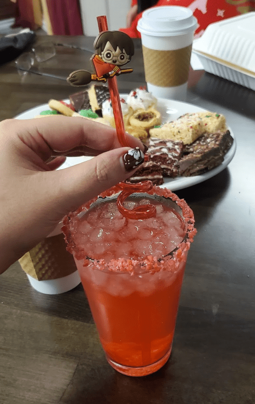 A hand holds a colorful drink with a character straw, surrounded by a plate of assorted desserts and coffee cups.