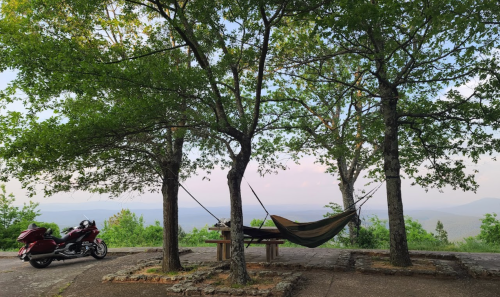 A hammock hangs between two trees with a motorcycle parked nearby, overlooking a scenic mountain view.