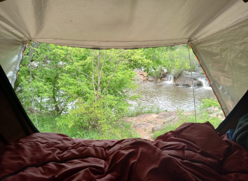 View from inside a tent, showcasing a river and lush greenery outside, with a cozy sleeping area in the foreground.