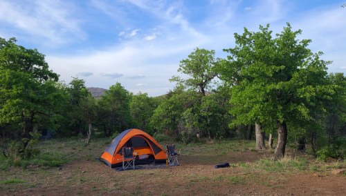 A bright orange tent set up in a green, wooded campsite with two folding chairs nearby under a blue sky.