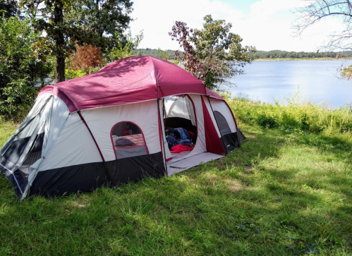 A large tent set up on grassy land by a lake, surrounded by trees and a clear blue sky.