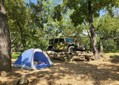 A blue tent set up near a green Jeep parked on rocks, surrounded by trees in a sunny outdoor setting.