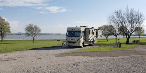 A parked RV by a lake, surrounded by grassy areas and trees under a clear blue sky.