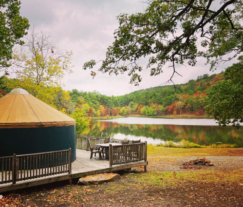 A yurt by a calm lake surrounded by trees with autumn foliage, under a cloudy sky.