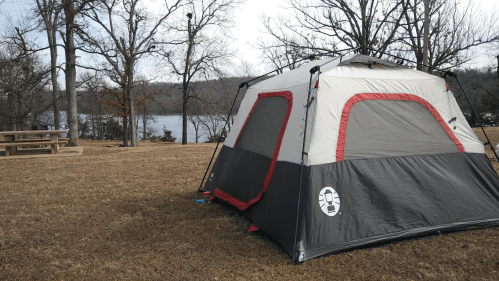A tent set up on grassy land near a lake, with trees in the background and a picnic table nearby.