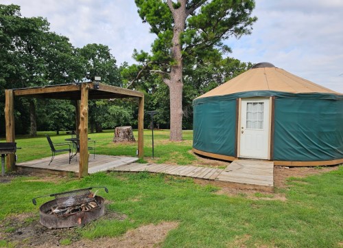 A yurt with a wooden deck and seating area, surrounded by grass and trees, with a fire pit in the foreground.