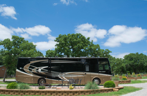 A parked RV in a scenic outdoor setting with trees and a blue sky, surrounded by gravel and a small seating area.