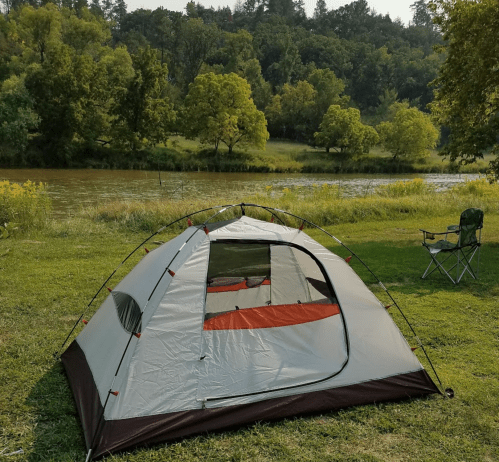 A tent set up on green grass by a river, surrounded by trees and a camping chair nearby.