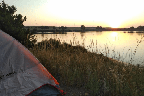 A tent by a calm lake at sunset, with tall grass and trees in the foreground.