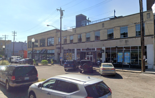 A street view of a commercial building with parked cars and pedestrians on a sunny day.