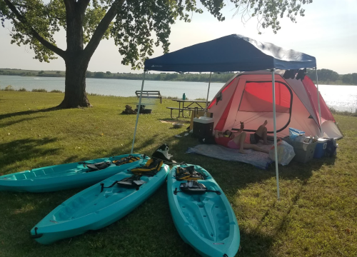 A campsite by a lake with three kayaks, a tent, and a picnic table under a tree on a sunny day.