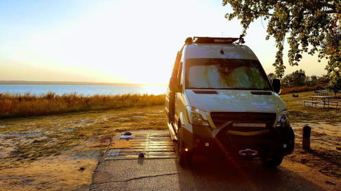 A camper van parked by a serene lake at sunset, with golden light reflecting on the water and trees in the background.