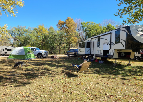 A campsite with a large RV, a green tent, a truck, and a person relaxing in a chair, surrounded by trees and grass.
