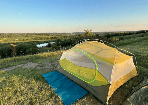 A tent set up on a grassy hill overlooking a river and distant landscape under a clear blue sky.