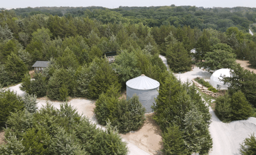 Aerial view of a wooded area with a silos and small buildings along winding paths. Lush greenery surrounds the scene.