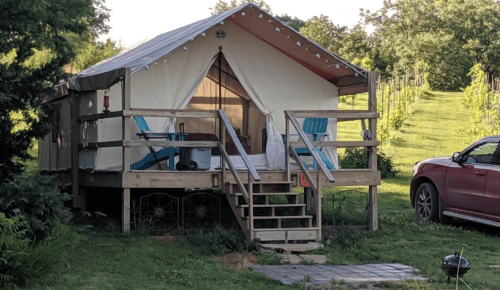 A cozy glamping tent on a wooden platform, surrounded by greenery and a car parked nearby.