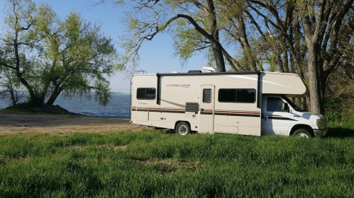 A parked RV near a lake, surrounded by green grass and trees, with a clear blue sky in the background.