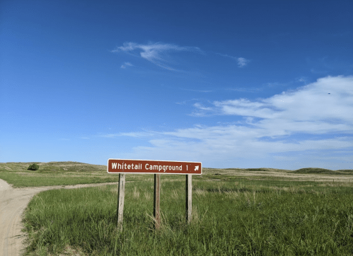 Sign for Whitetail Campground 1, surrounded by grassy fields under a clear blue sky.