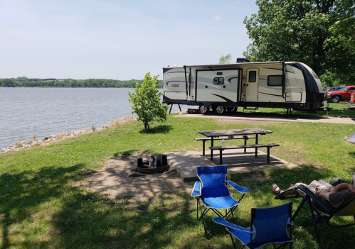 A camper parked by a lake, with a picnic table, fire pit, and blue chairs on a grassy area under a clear sky.