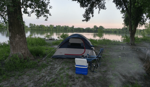 A tent set up by a calm river, with a cooler and camping chair nearby, surrounded by trees and greenery.