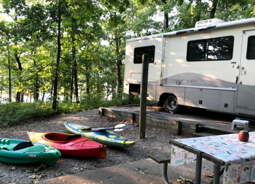 A campsite with an RV, colorful kayaks, and a picnic table surrounded by trees near a lake.