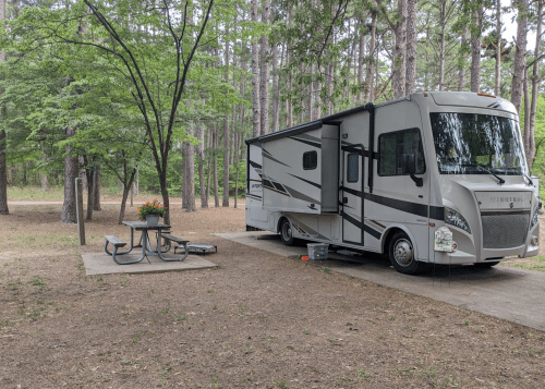 A parked RV beside a picnic table in a wooded area with trees and a gravel path.