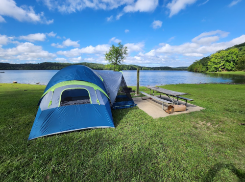 A blue and green tent by a lake, with a picnic table nearby and trees in the background under a partly cloudy sky.