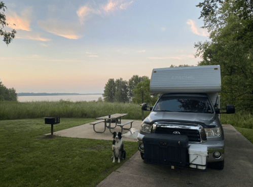 A truck camper parked by a lake at sunset, with a dog sitting nearby and a picnic table in the foreground.