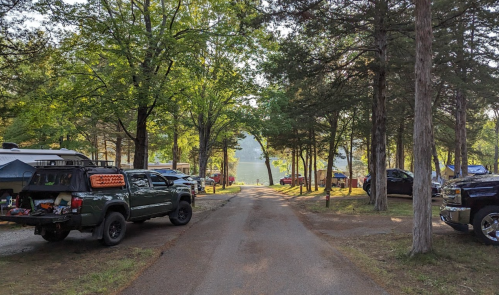A tree-lined campsite road with parked vehicles and a view of a lake in the distance.