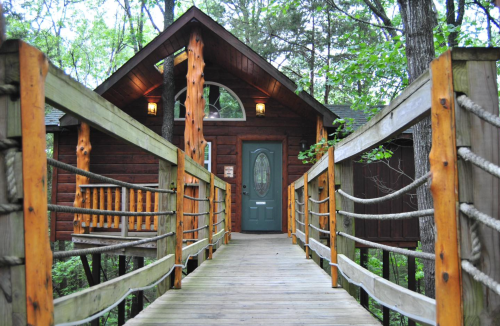 A wooden walkway leads to a rustic cabin surrounded by trees, featuring a green door and warm lighting.