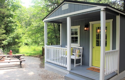A cozy blue cabin with a yellow door and porch, surrounded by greenery and a picnic area nearby.
