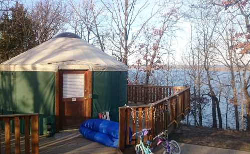 A yurt on a wooden deck overlooking a lake, surrounded by trees, with a blue inflatable mattress and bicycles nearby.