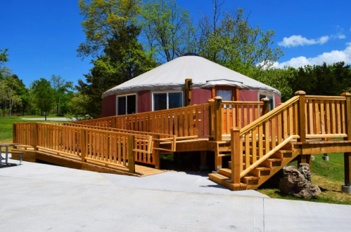 A wooden deck with ramps leads to a round yurt surrounded by greenery and blue skies.