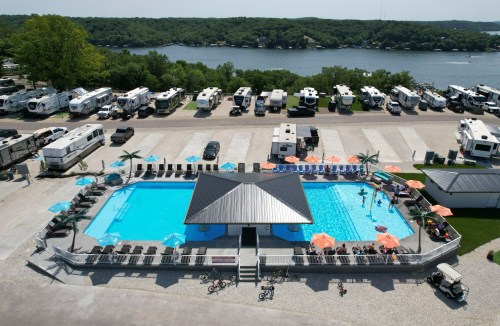 Aerial view of a pool area with umbrellas, surrounded by RVs and a lake in the background.