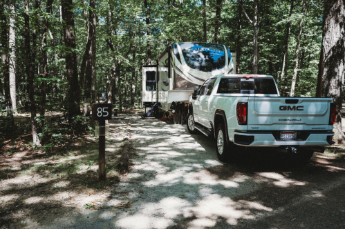 A white GMC truck parked next to a large RV in a wooded campsite, with a sign displaying the number 85.