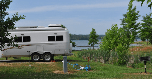 A silver camper parked near a lake, surrounded by greenery and trees on a sunny day.