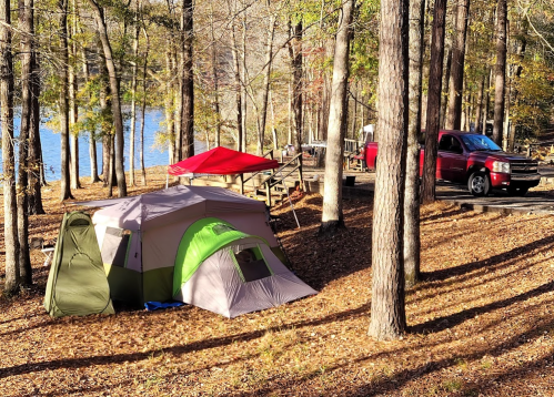 A campsite in the woods with two tents, a red canopy, and a truck parked nearby, beside a lake.