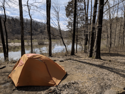 A bright orange tent set up by a river, surrounded by bare trees and a scenic landscape.