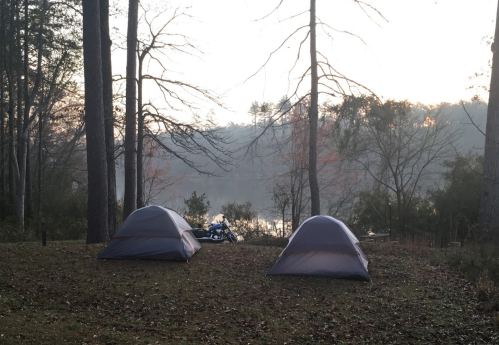 Two tents set up near a calm lake, surrounded by trees, with a motorcycle parked in the background at dawn.