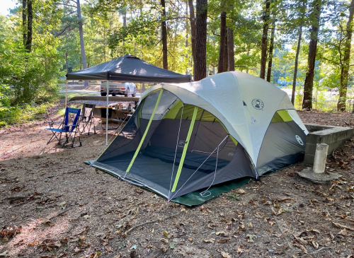 A spacious tent set up in a wooded campsite, with a canopy and picnic area nearby.