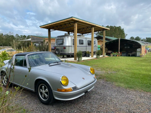 A silver convertible car parked in front of a modern shelter with an Airstream trailer and greenery in the background.