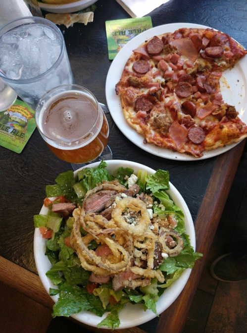 A table with a salad topped with fried onions, a slice of pizza with pepperoni and ham, and a glass of beer.